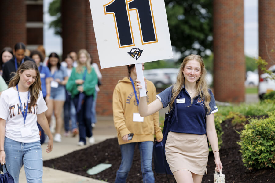 Student in navy polo holding large number 11 sign, walking outside campus building with group of peers.