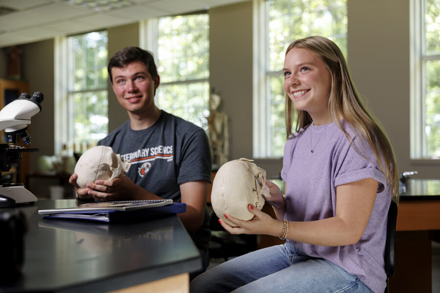 Two students in a science lab, smiling and holding anatomical skull models near a microscope and windows.