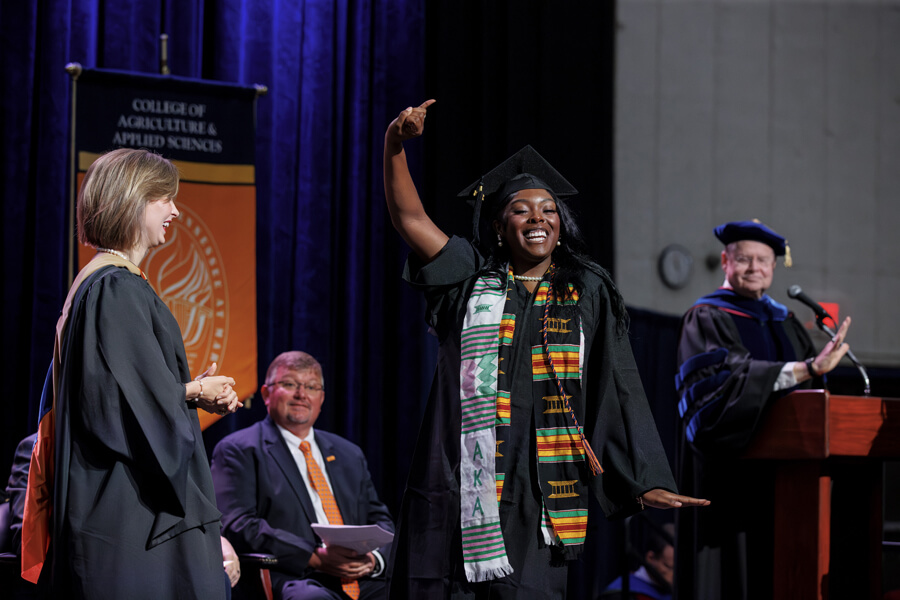 Joyful graduate in cap and kente stole celebrates on stage during College of Agriculture & Applied Sciences commencement ceremony.