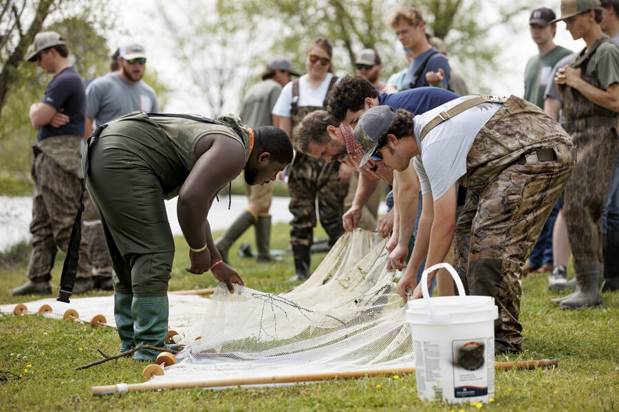 Group of people in military and casual attire examining a fishing net with equipment on grassy ground near trees.
