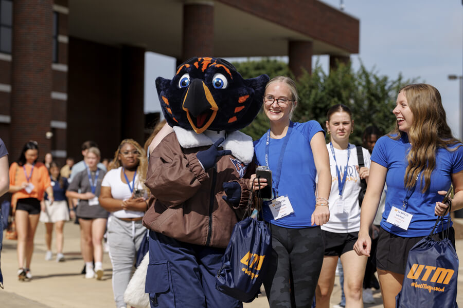 University mascot with students in blue shirts walking together on campus on a sunny day