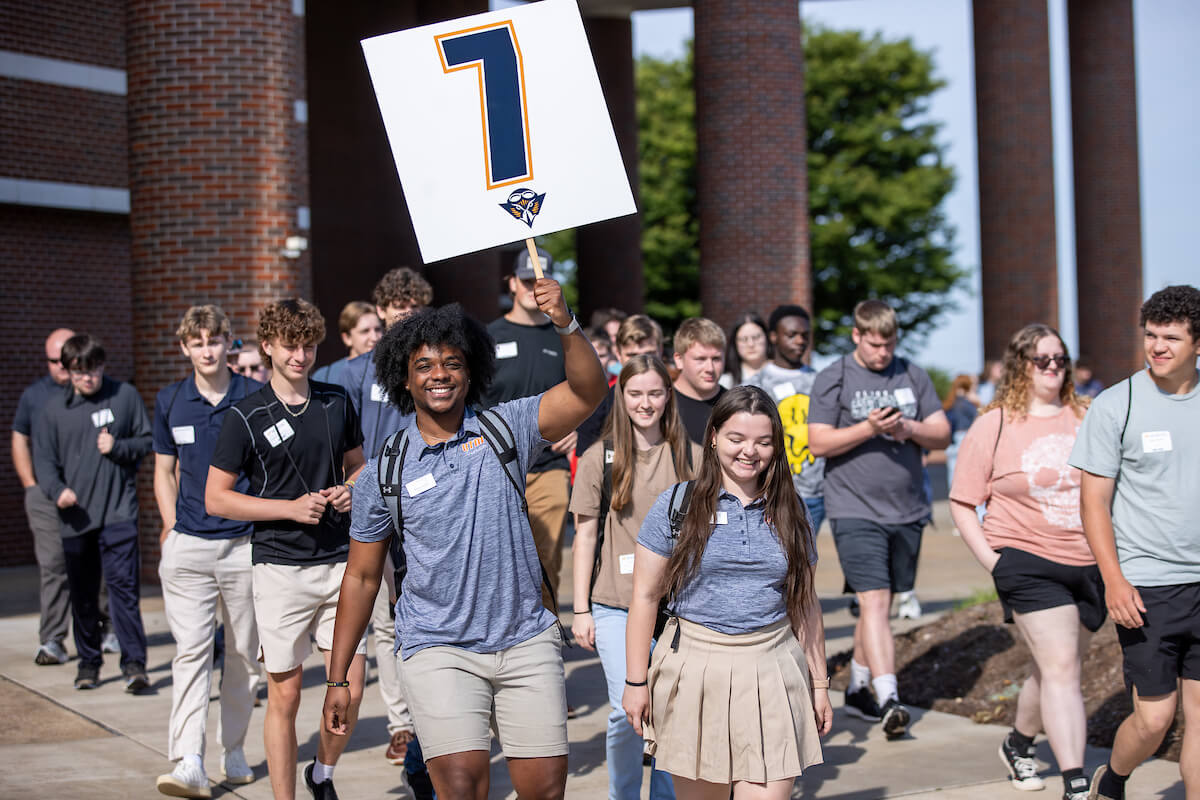A tour leader carries a sign with the group number seven as he leads his group across the UTM quad.